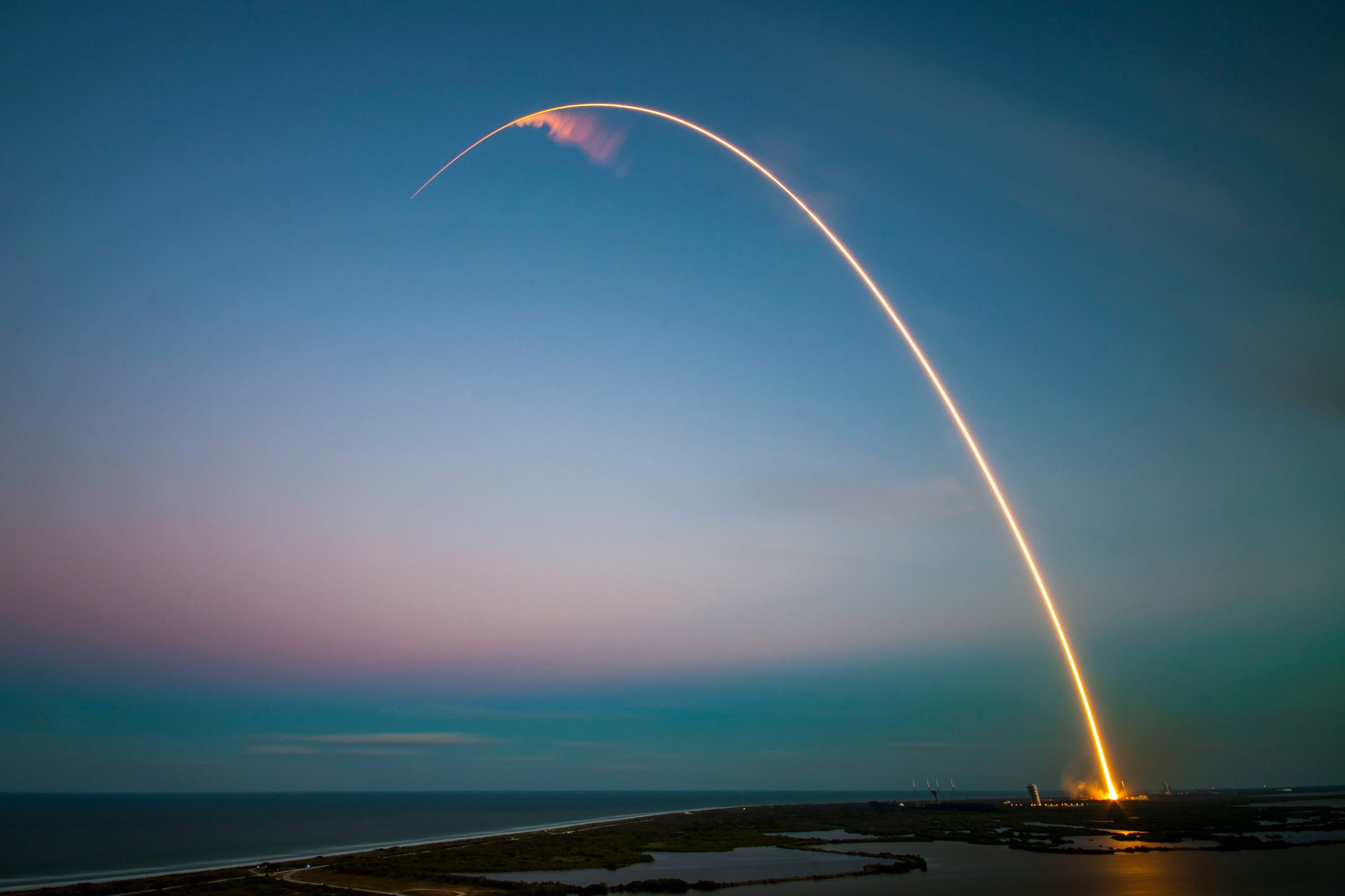 Long-exposure rocket trail arcing across a twilight sky over the coast